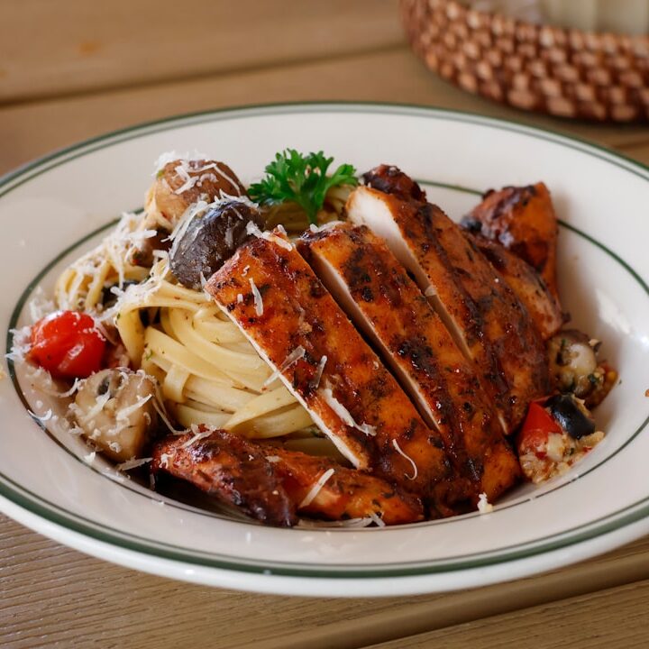 Sliced grilled chicken breast over pasta with vegetables.