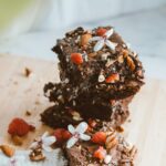 Stack of chocolate brownies garnished with nuts, berries, and flowers on a wooden board.
